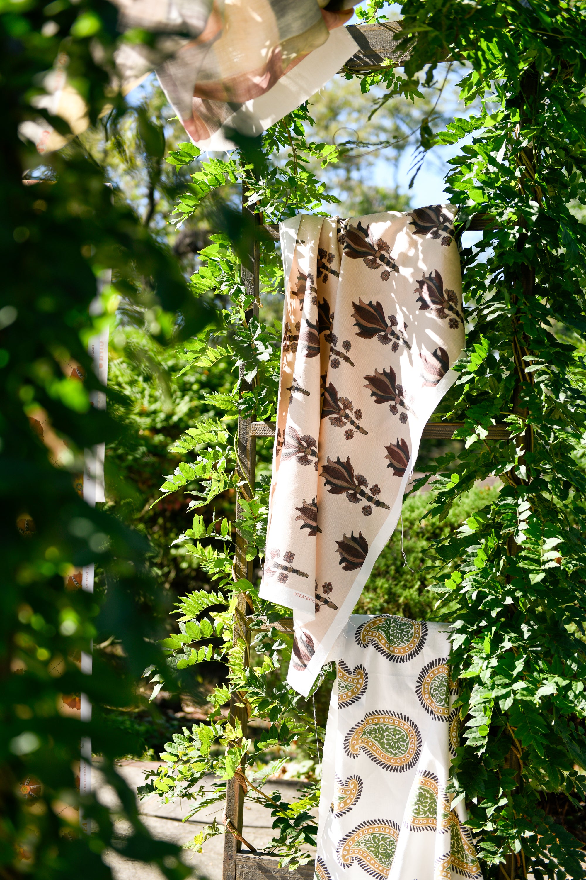 A yard of fabric with a floral pattern hanging outdoors among greenery.