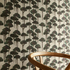 A section of wallpaper with a pattern of umbrella pines in various shades of gray and green on a light background, with part of a wooden chair visible in the frame.
