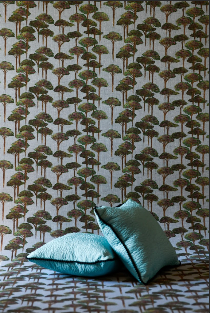 A room with wallpaper featuring a repeating pattern of umbrella-shaped pine trees in various shades of green, beige, and brown tones. Two pillows with a solid blue texture are placed in the foreground.

